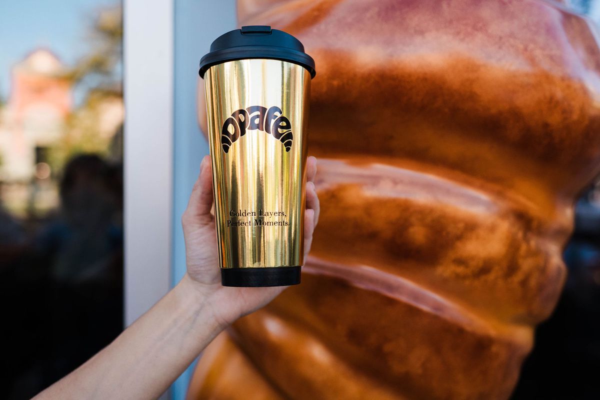 Gold Opafe tumbler held up beside an oversized croissant